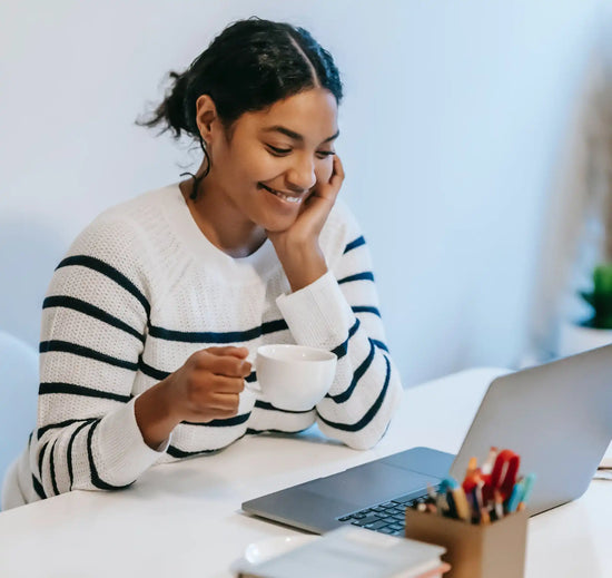 A person in a striped sweater smiling while holding a coffee cup at a desk with a laptop.